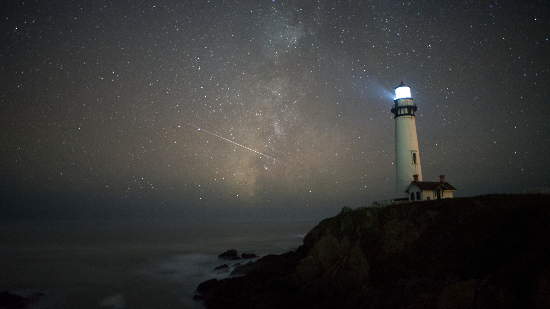 A glowing light house stands on the edge of a coastline where above it a meteor streaks in the night sky