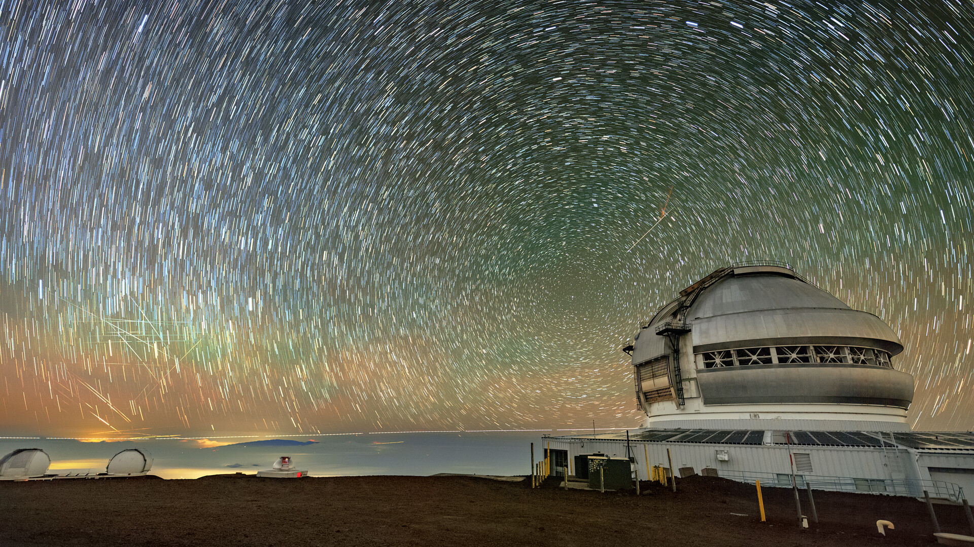 In this long exposure image, a series of rings and streaks in concentric circles fill the colorful blue, orange, and green night sky with a domed roof of an observatory below.