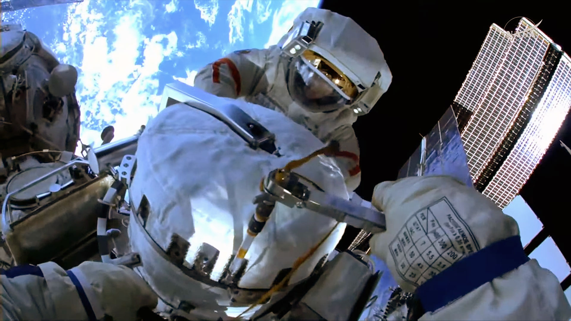 a camera mounted on a cosmonaut&#039;s spacesuit helmet captures a first-person view of another cosmonaut and Earth behind him while outside a space station.