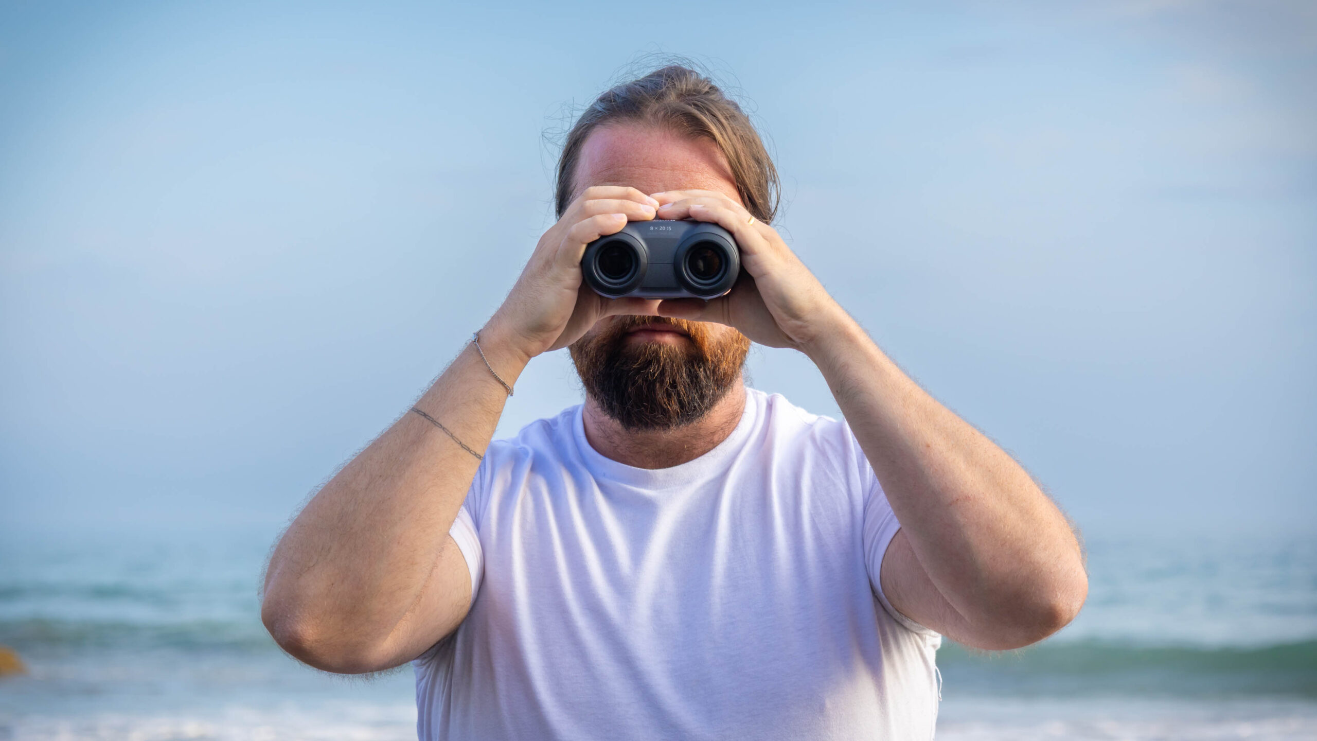 A male using the Canon 8x20 IS binoculars, looking directly at the camera, with the sea behind them.