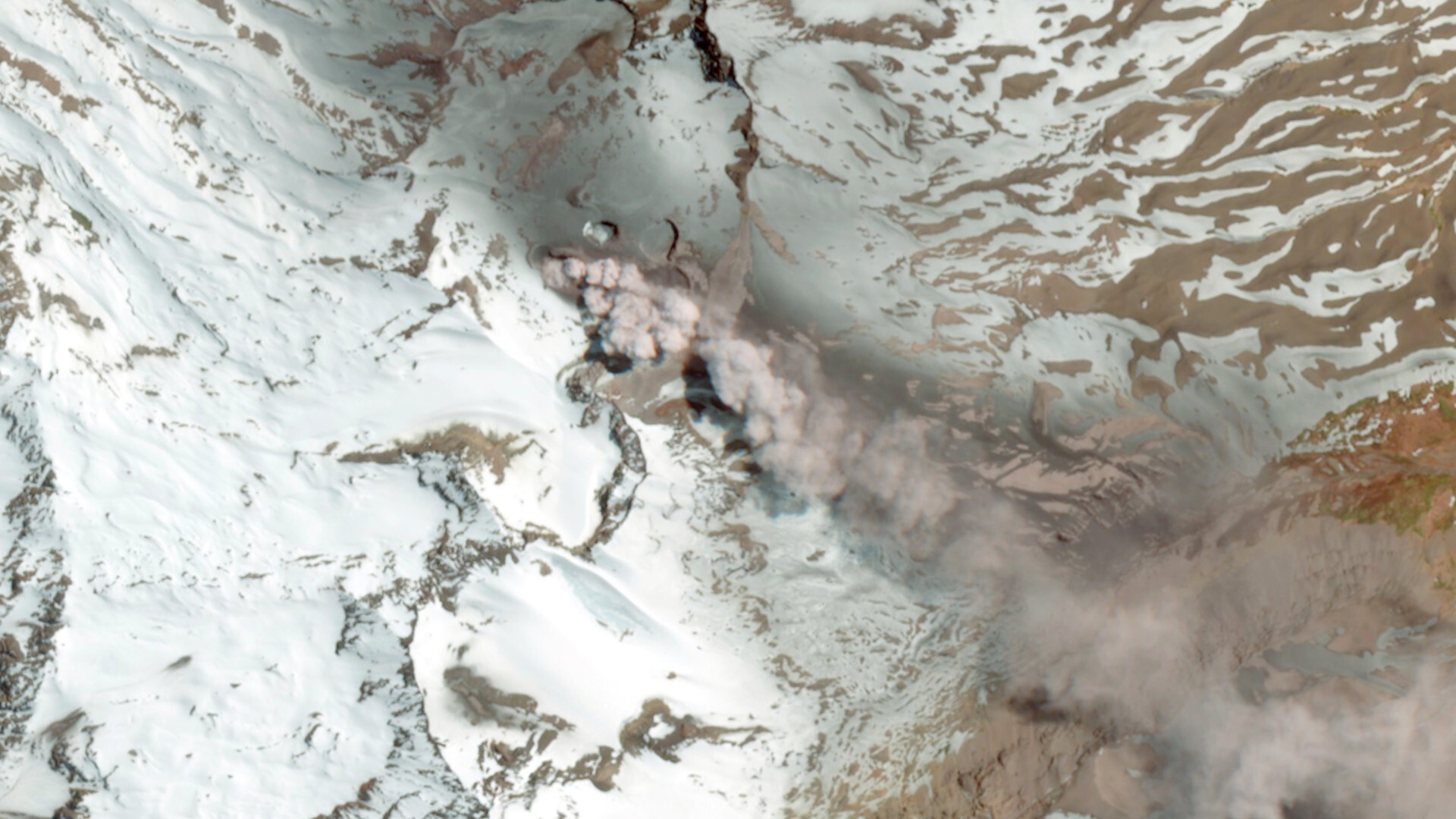 An image from space showing a gray plume of smoke drifting over brown and snowy mountains