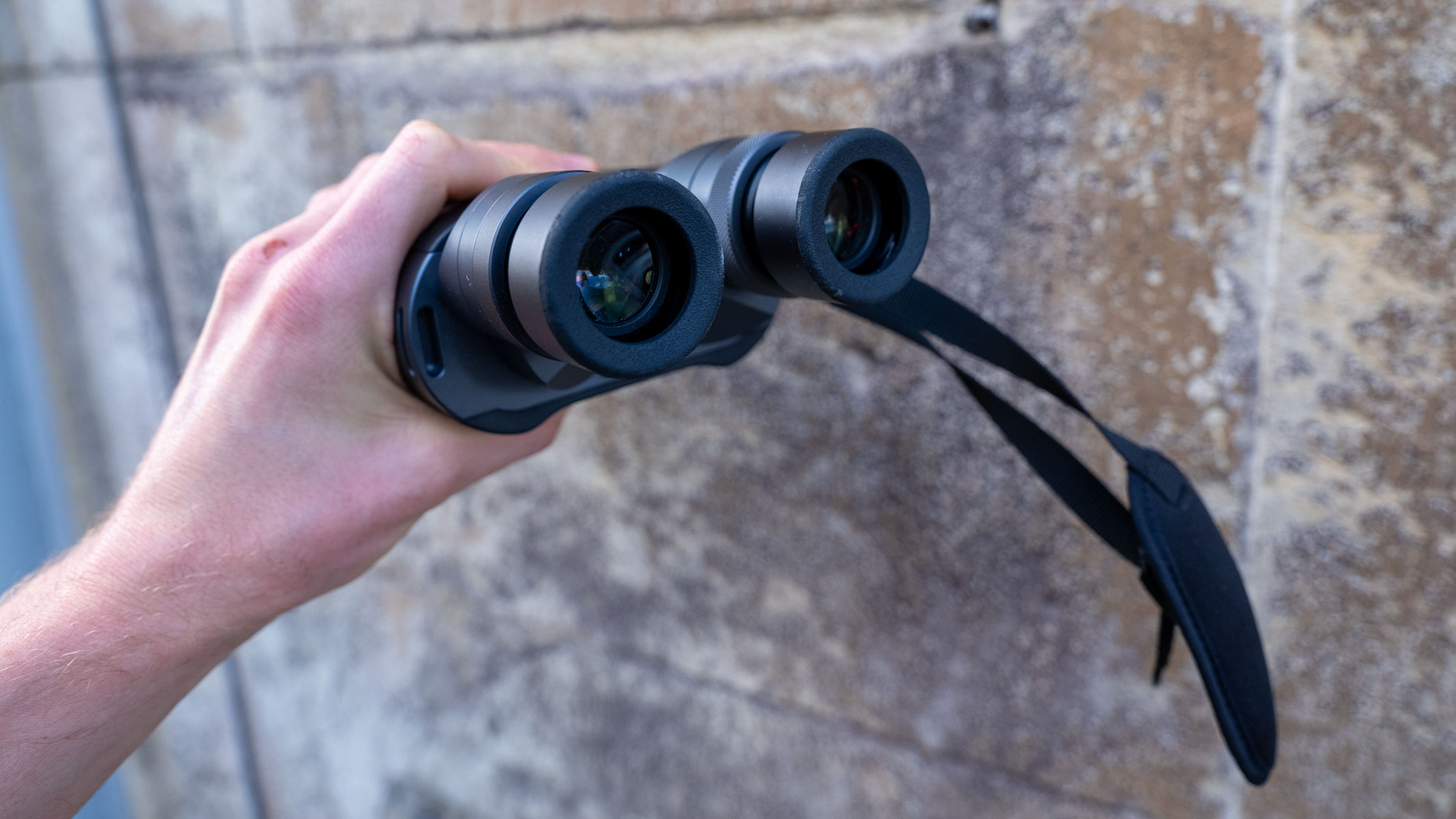 A hand holding the Fujifilm Techno-Stabi TS-L 1640 image-stabilized binoculars with a beige wall in the background.
