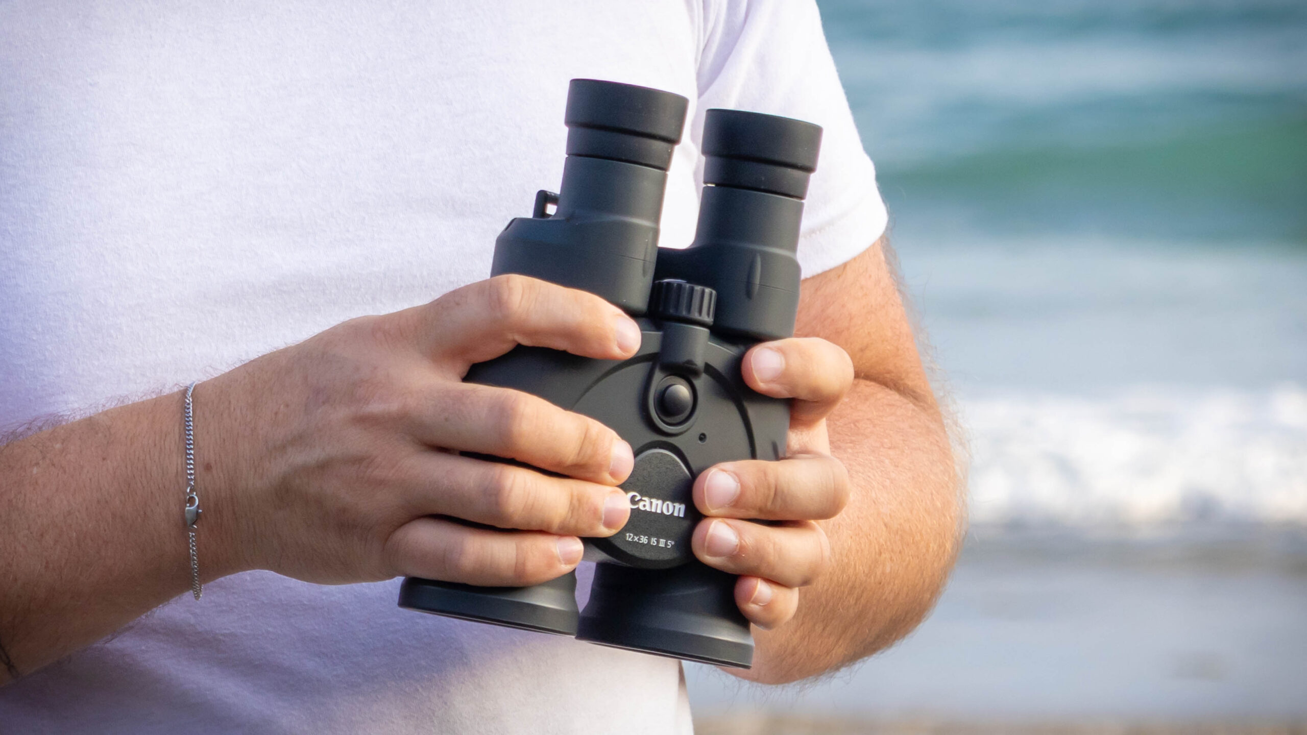 A close up of the Canon 12x36 IS binoculars being held by someone, next to the sea.