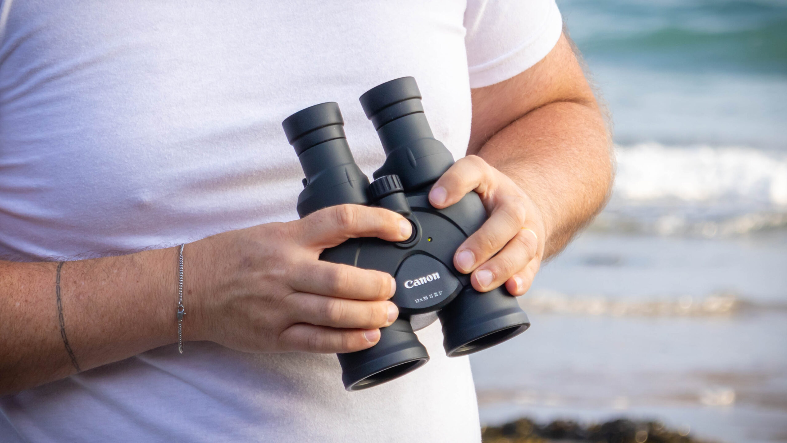 A person holding the IS button down on the Canon 12x36 IS binoculars, with the sea behind them.