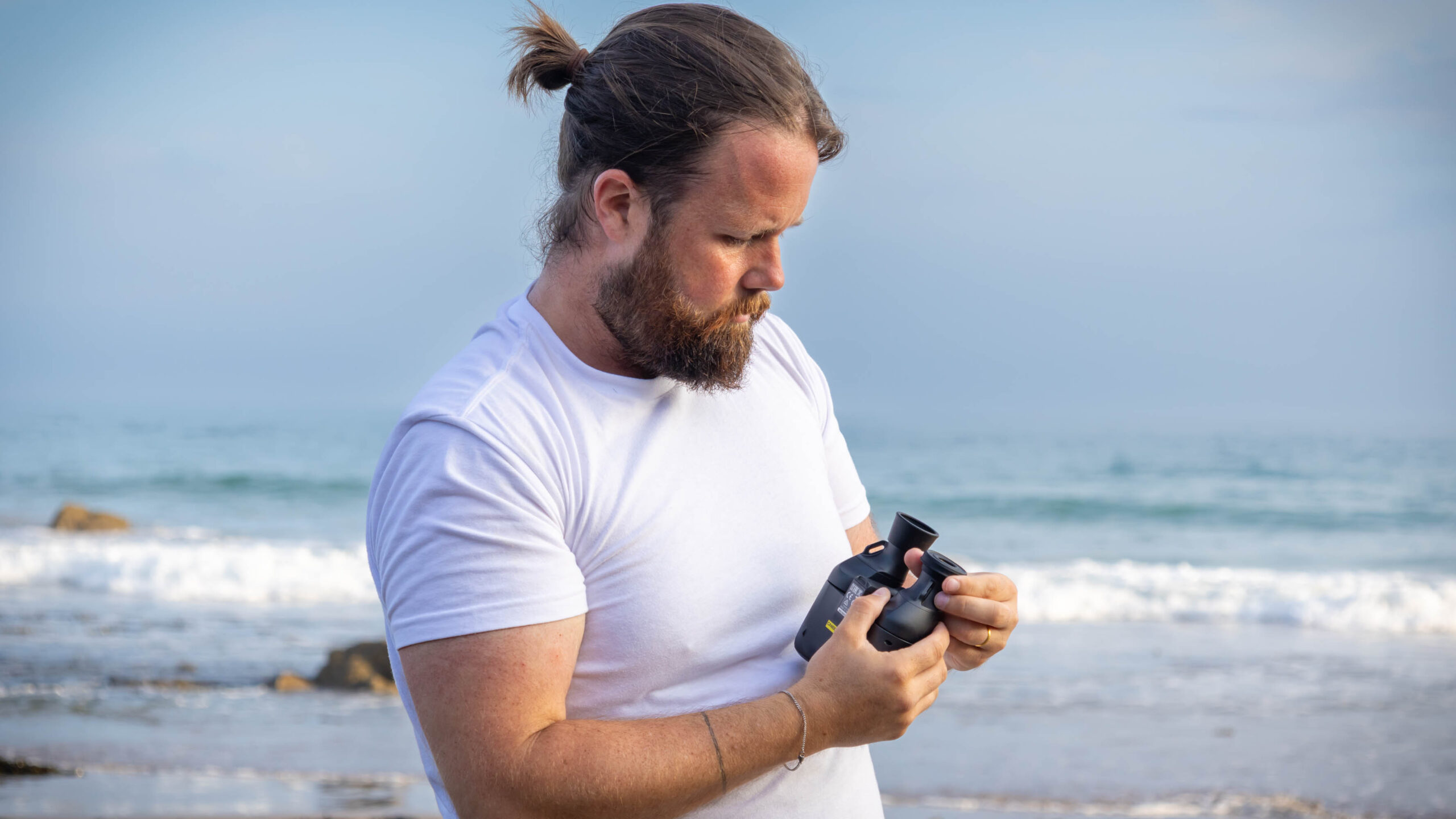 A male holding the Canon 8x20 IS binoculars, twisting the eyecups, with the sea behind them.