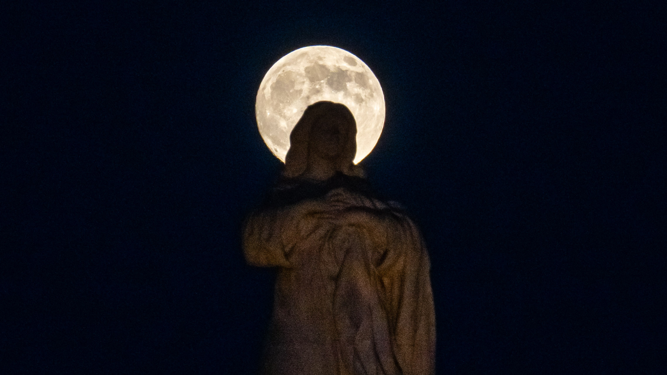 A full moon is pictured rising behind a stone statue depicting the Virgin Mary, giving the appearance of a halo shining around its head.
