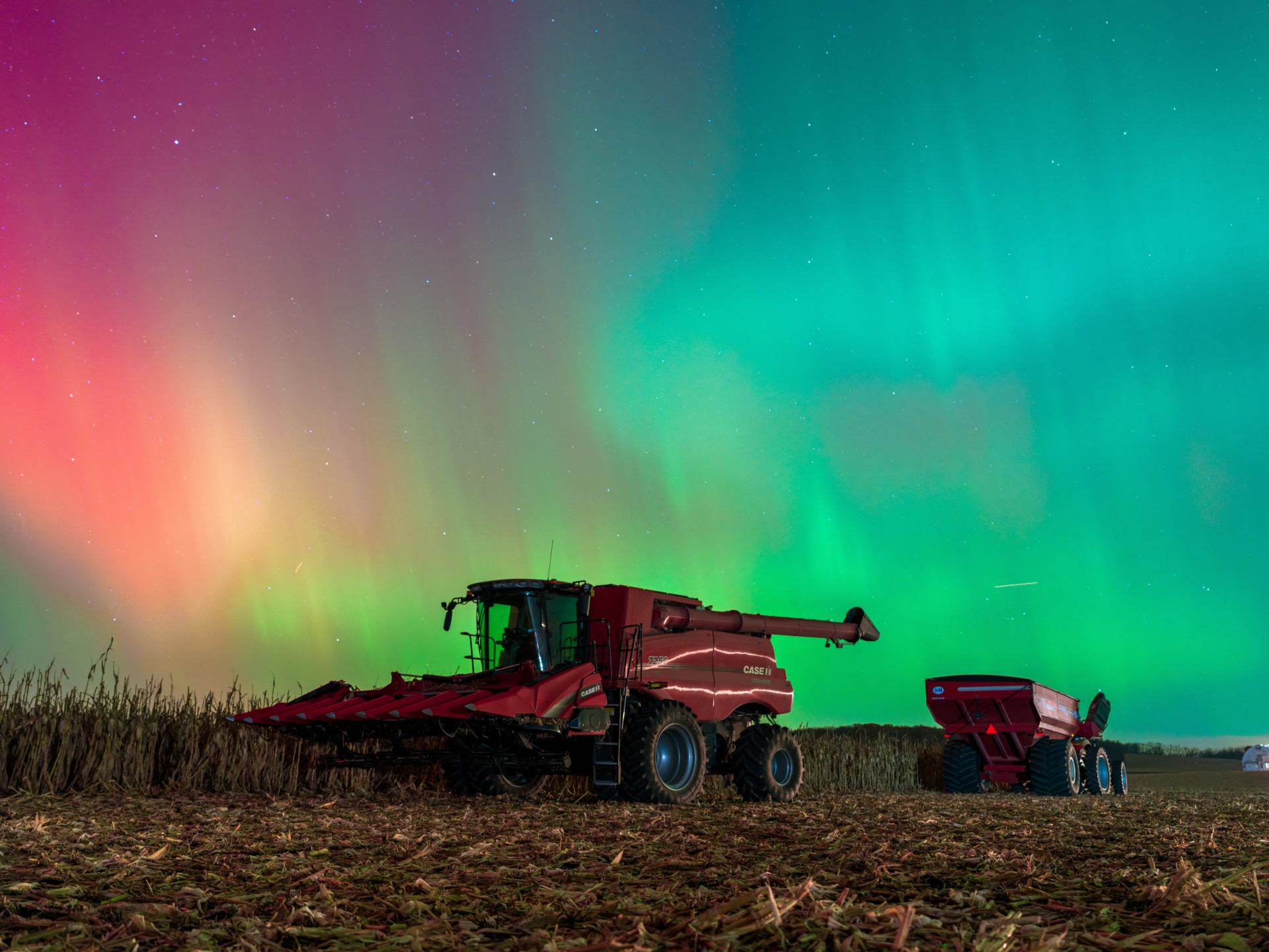 red, magenta and green auroras appear as curtains of light in the sky above farming equipment in a field.