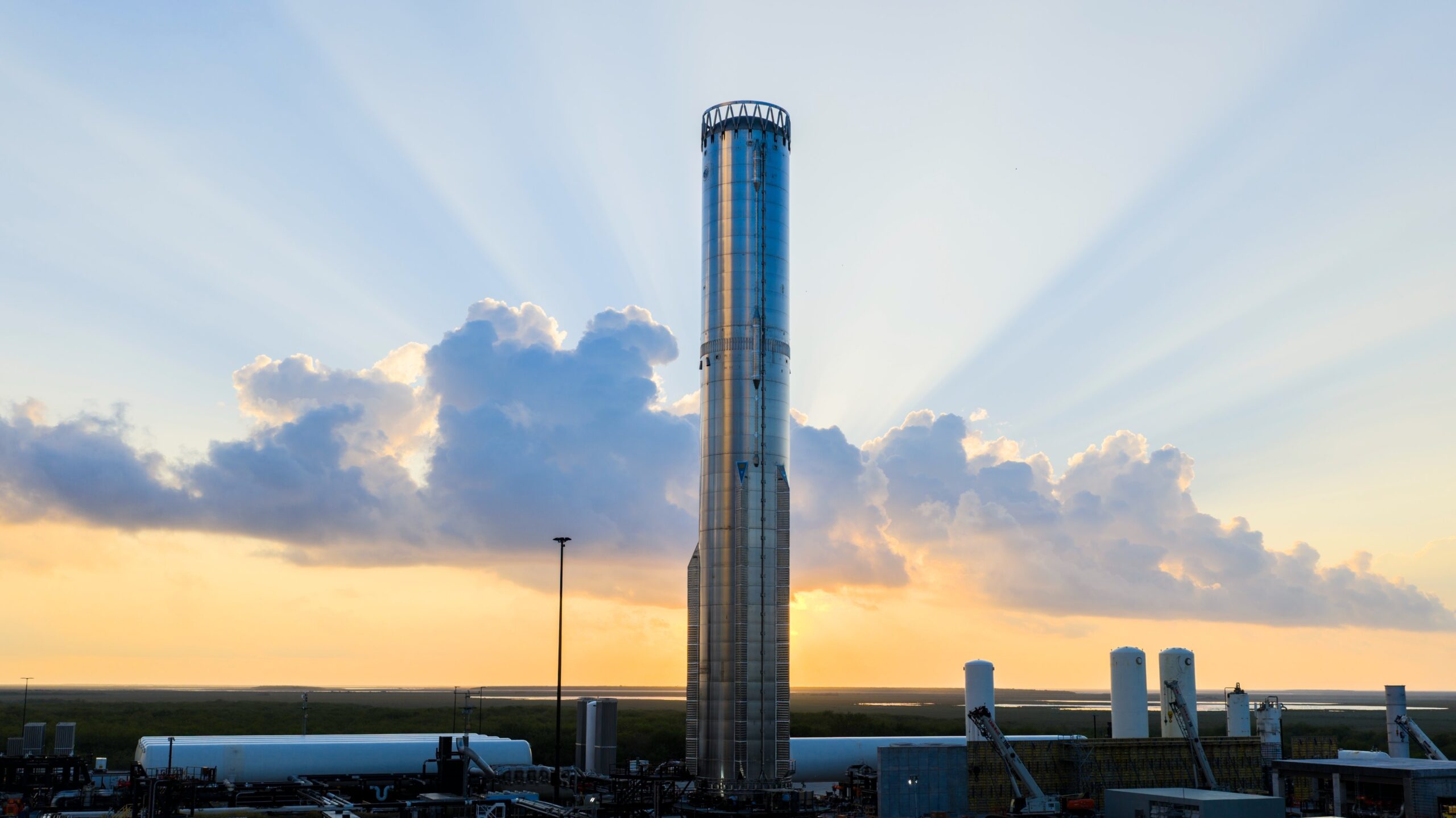 A tall chrome cylinder stands in front of a setting sun and protruding light rays.