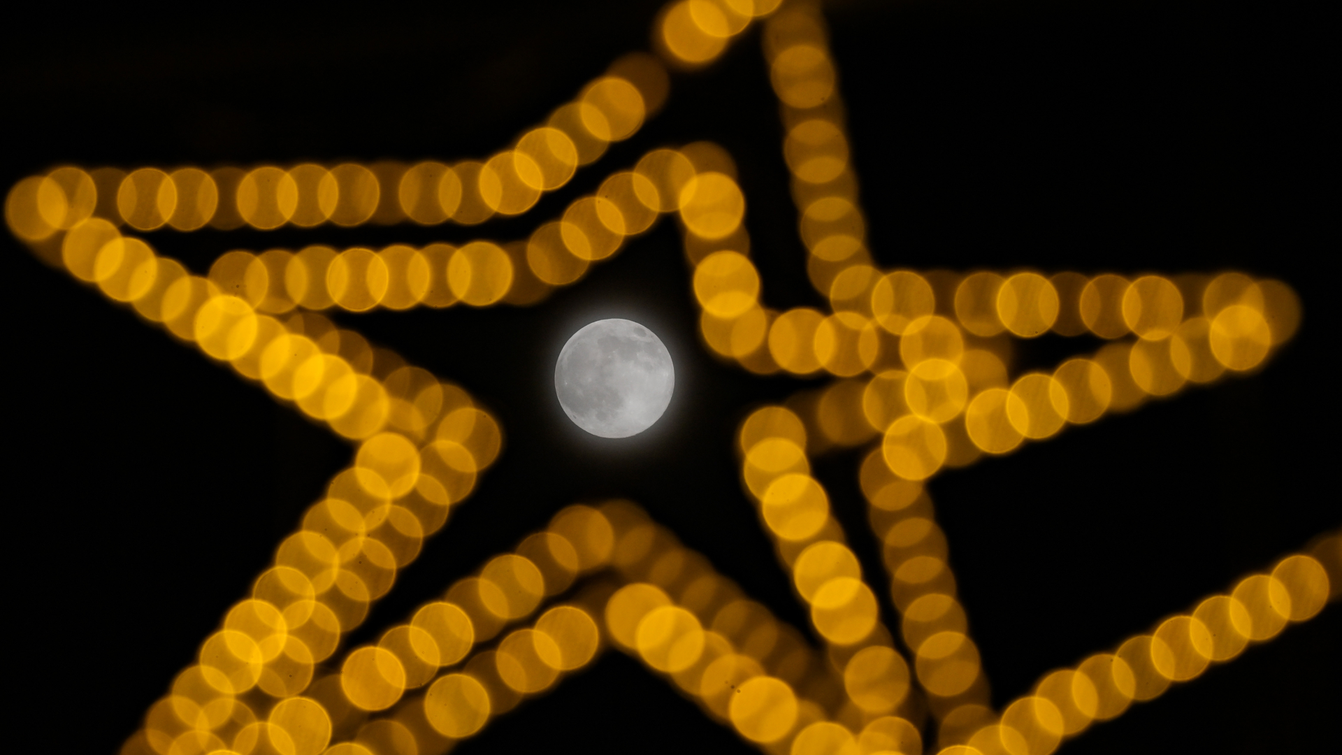 A full moon is pictured shining in the middle of concentric yellow stars made from artificial lights against a black sky.