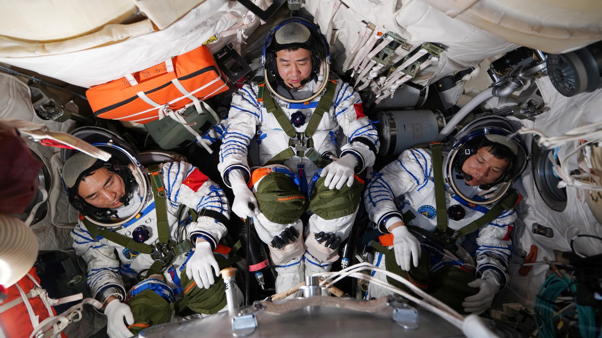 three astronauts in white spacesuits inside a cramped capsule full of equipment strapped to bulkheads