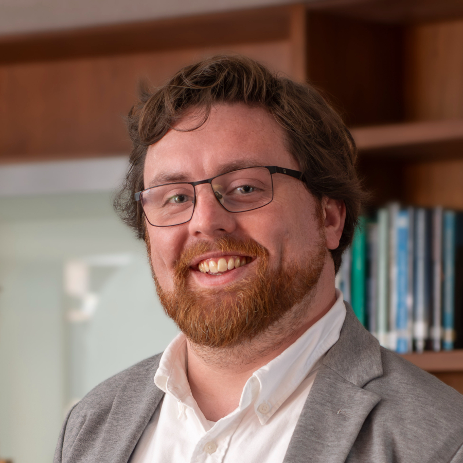 A man wearing glasses with red hair and a beard wearing a gray suit and white shirt looks at the camera