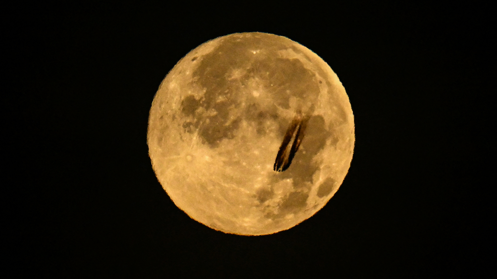 An image of a pale yellow full moon pictured against a black sky, with the dark plume of a passing commercial airliner darkening the lower right portion of its ancient surface. Bright craters can also be seen contrasting with dark lunar seas.