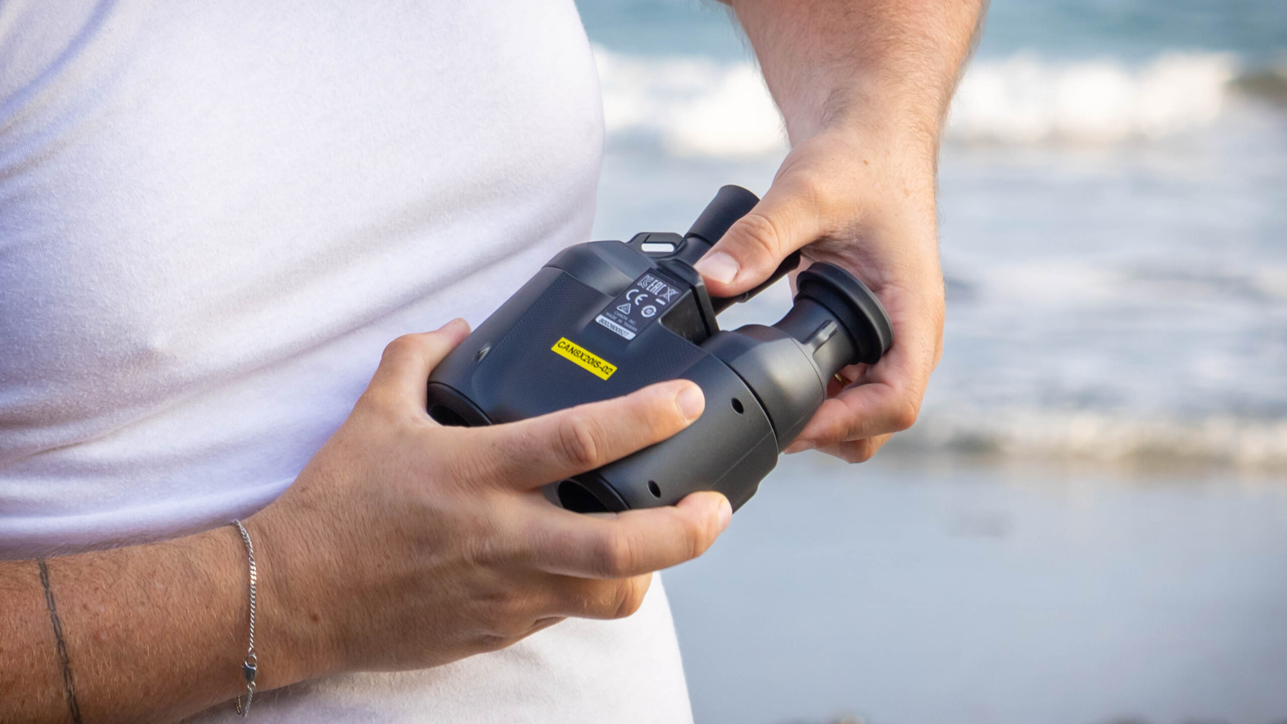 A person showing the back of the Canon 8x20 IS binoculars, with the sea behind them.