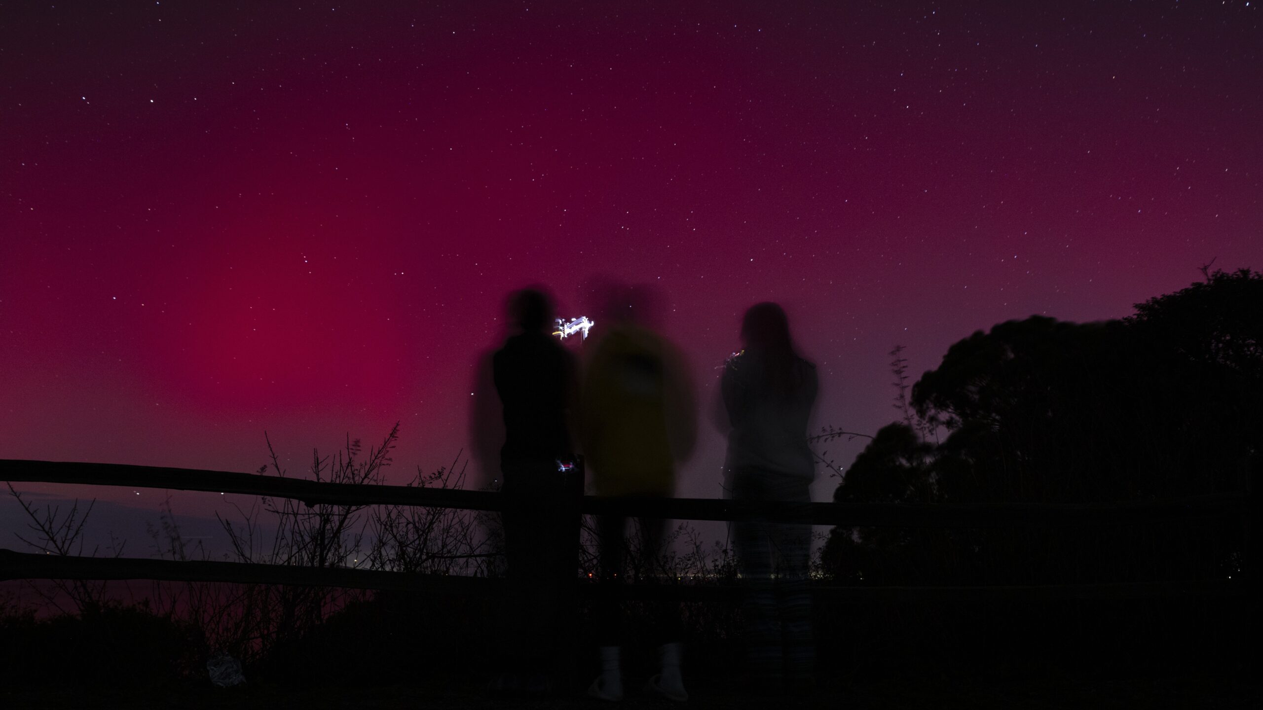 three people stand looking at at a red sky filled with aurora