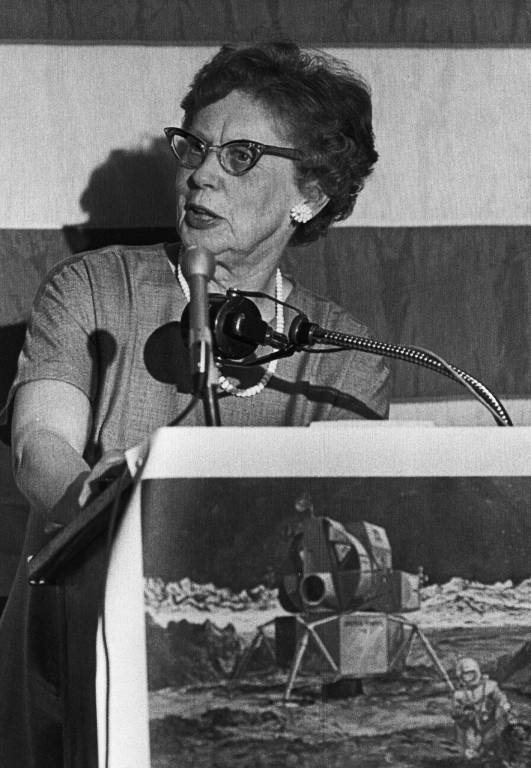 a woman speaks into a microphone at a lectern in front of the american flag