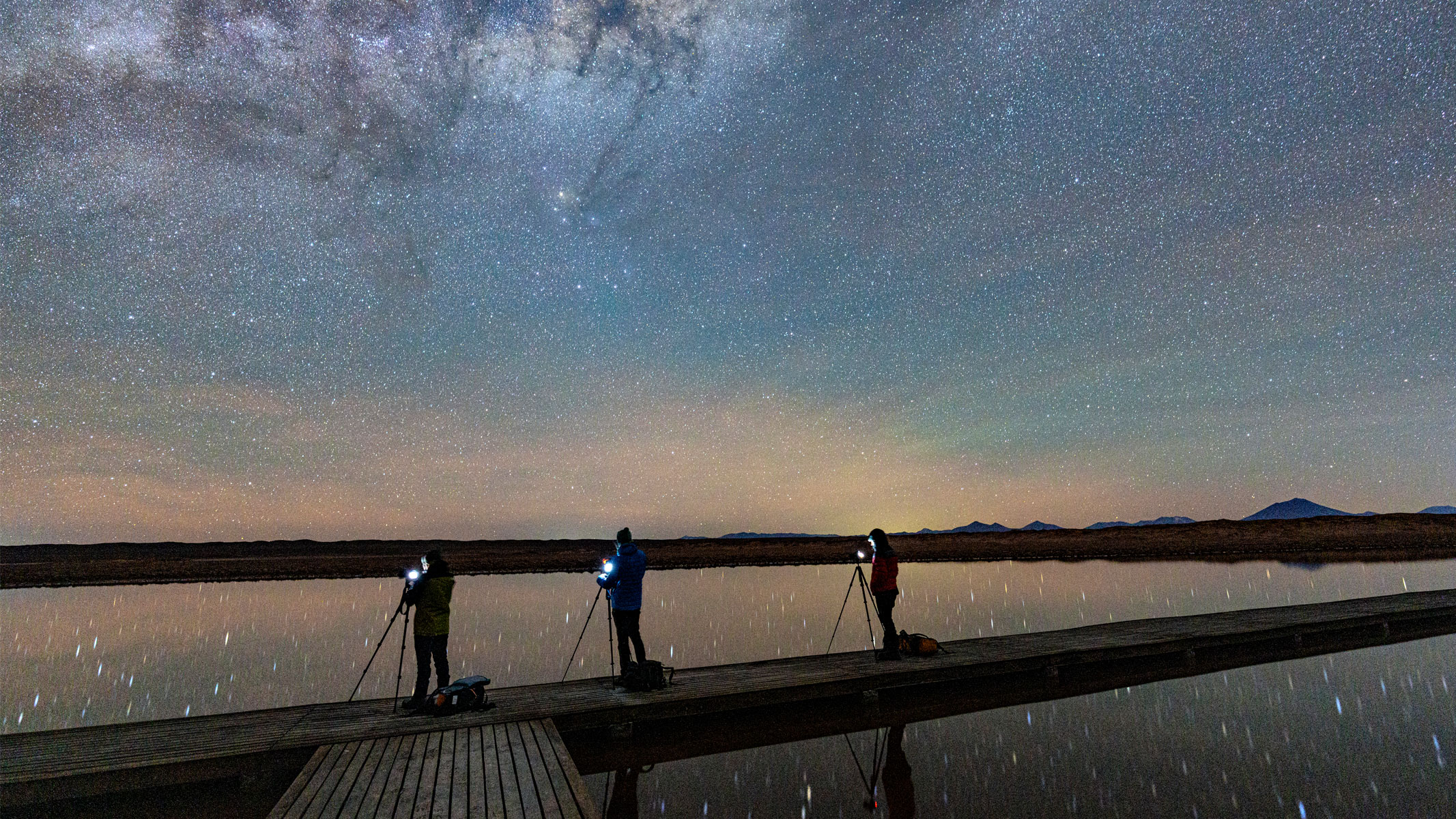 Three people stood near water with cameras with a twilight sky in front of them.