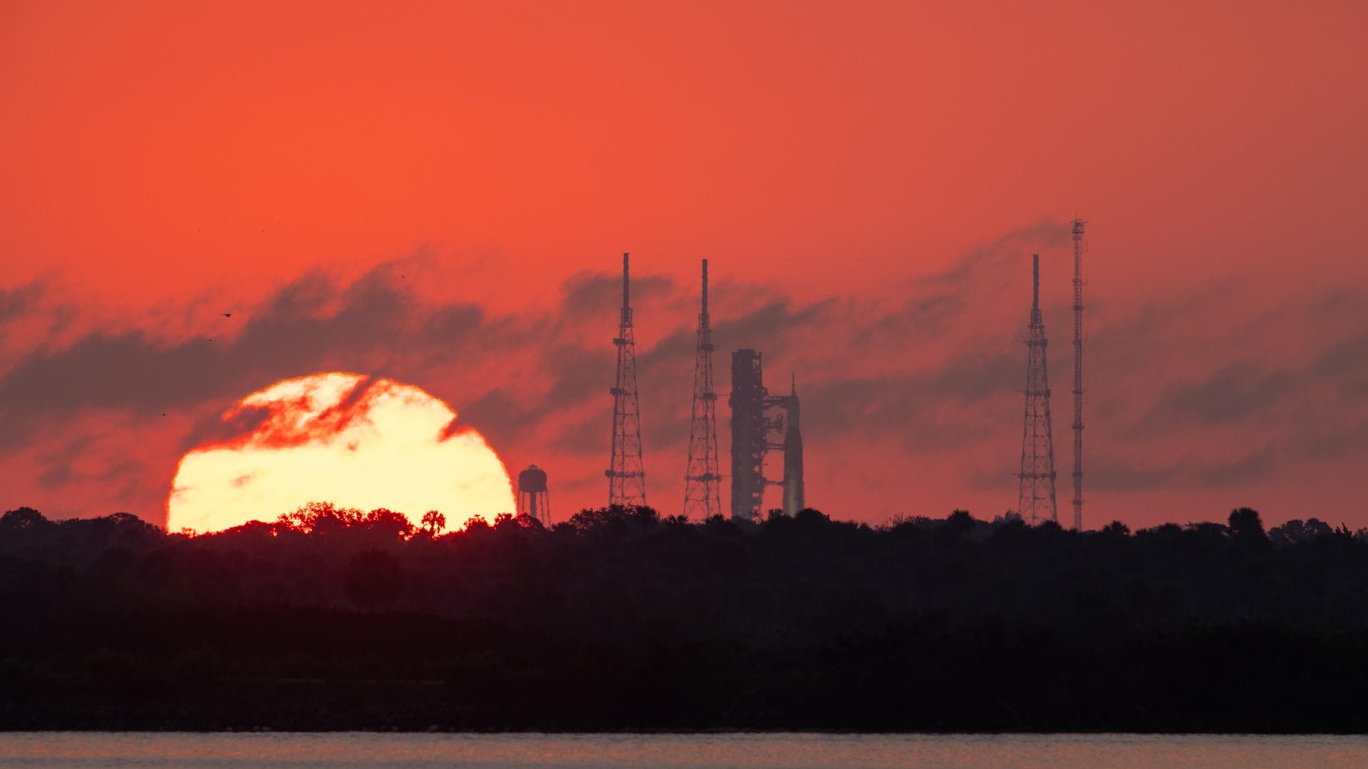 a massive-looking sun rises in a red sky behind the silhouettes of several skeletal metal towers and a rocket upright on a launch pad