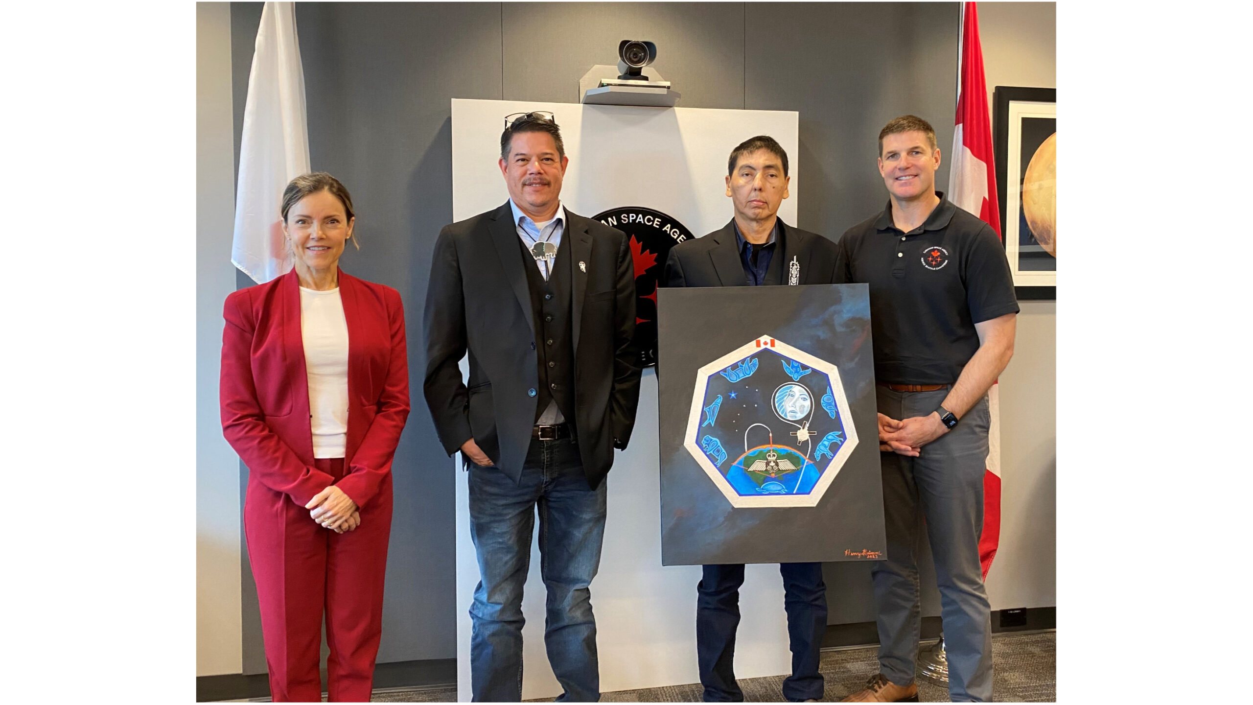 three men and one woman stand inside a room. one of the men is holding a painting of a space mission patch