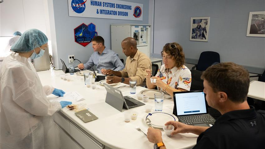 four astronauts in street clothes sit at a white table, trying out space foods
