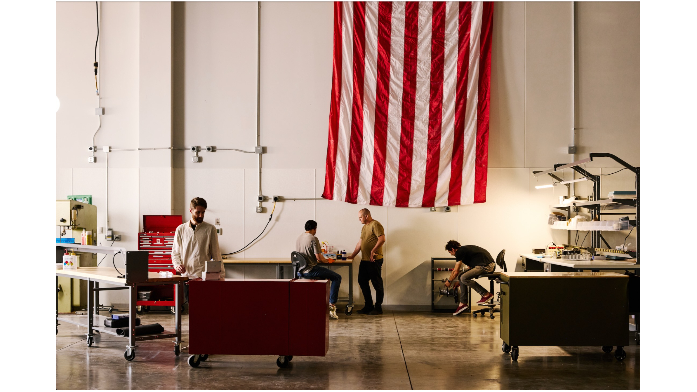 four men work inside a large, white-walled room with an american flag in the background