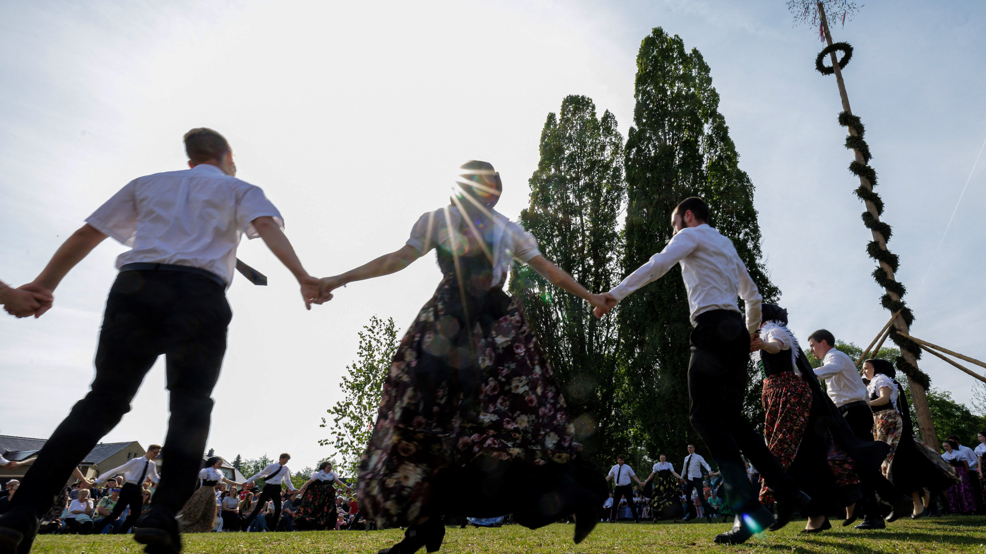 People are photographed holding hands and dancing close to a Maypole with tall trees in the background.