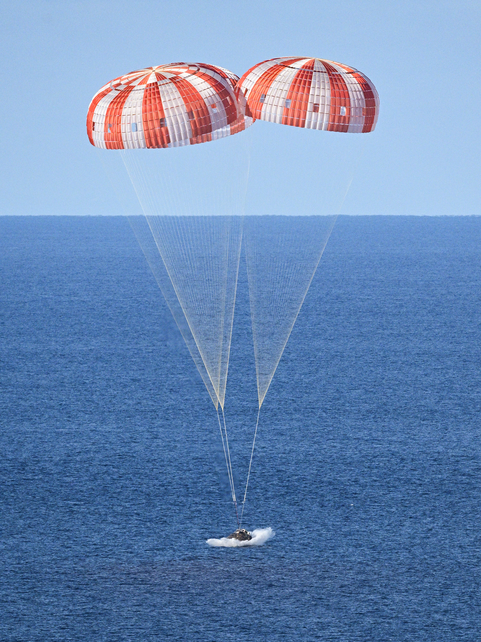 A space capsule splashes down in a blue ocean descending under three large parachutes.