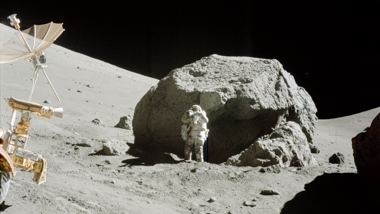 An astronaut in a white spacesuit stands next to a large gray boulder on the Moon with a small rover in front