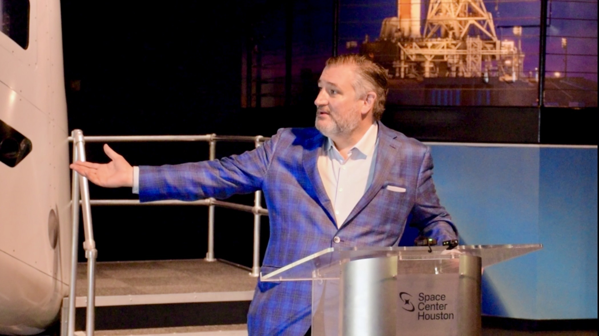 A man in a blue and black checkered plaid jacket stands at a podium and extends his arm to the left.