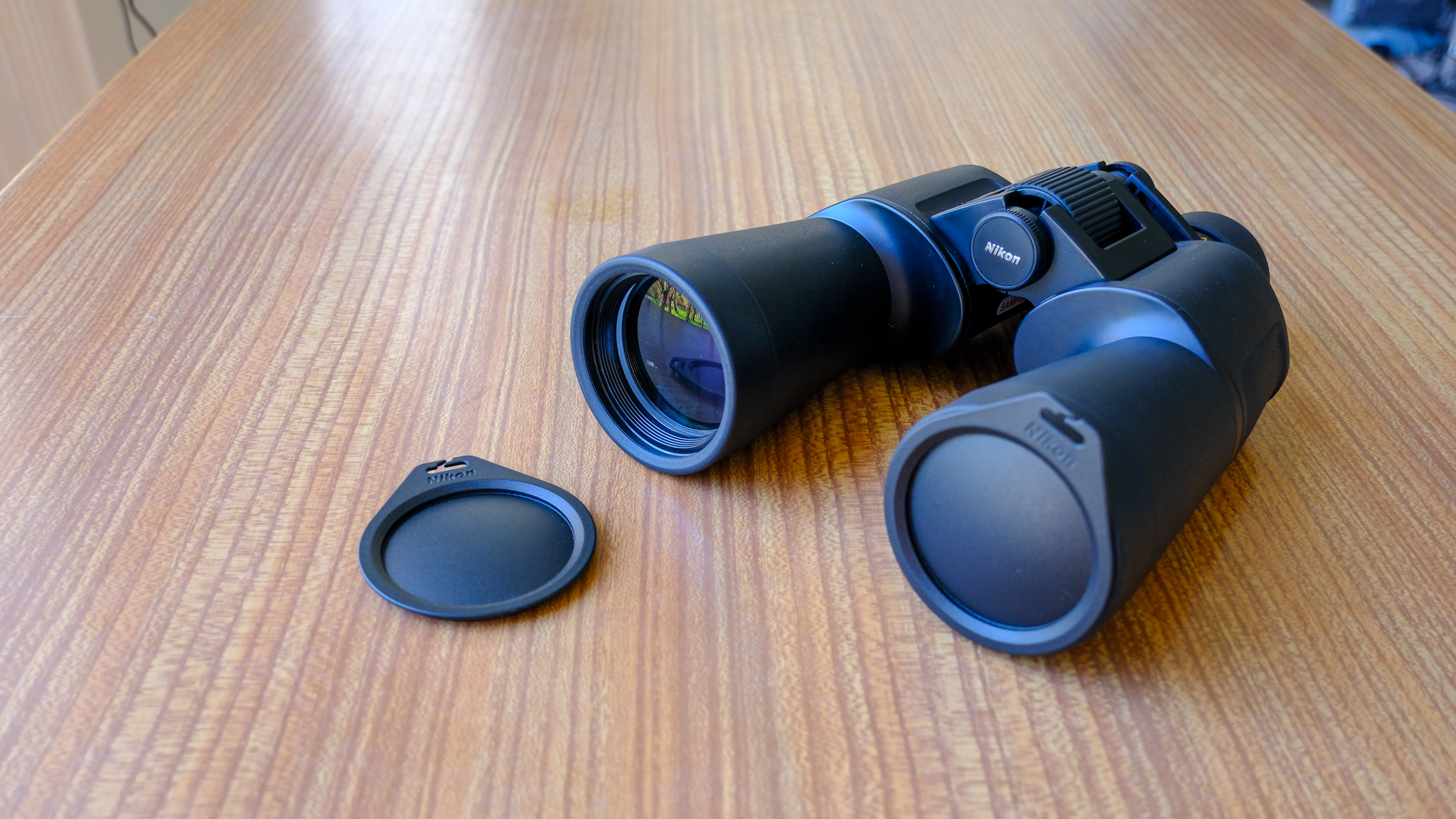 The Nikon Action 7x50 binoculars on a wooden table, with one lens cap on and one lens cap on the table.